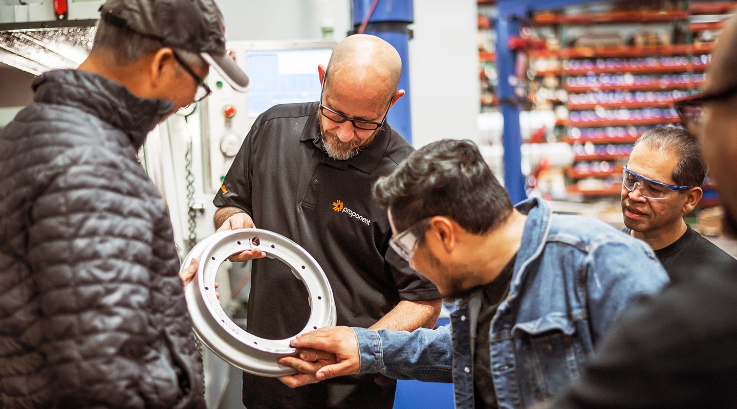 A man holds a part, while showing it to four people. One man is feeling the part and inspecting it