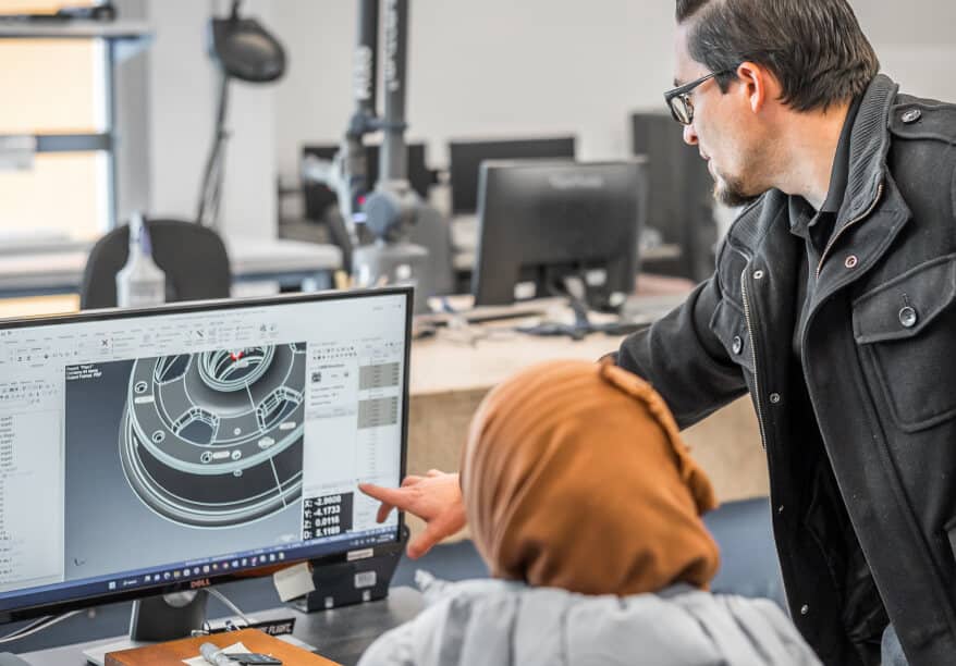 A man in a black jacket, points to a CAD design on a screen, with someone following along