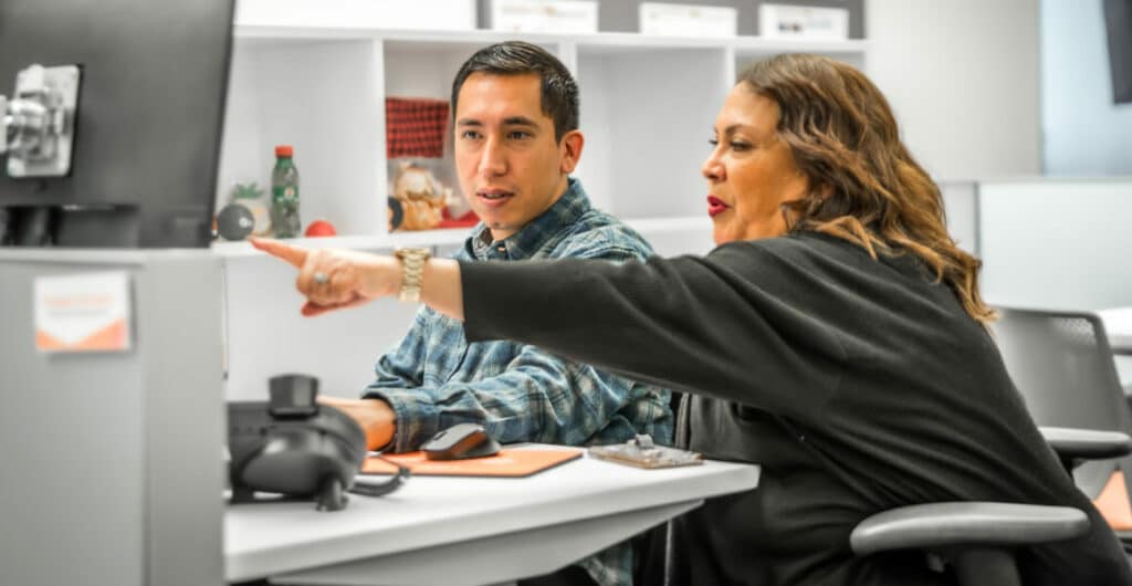 A woman in a dark grey top points to a computer screen with a man in a plaid shirt, following along