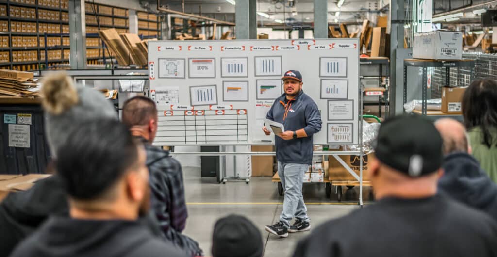 A man speaks to his team, with charts on a whiteboard behind him