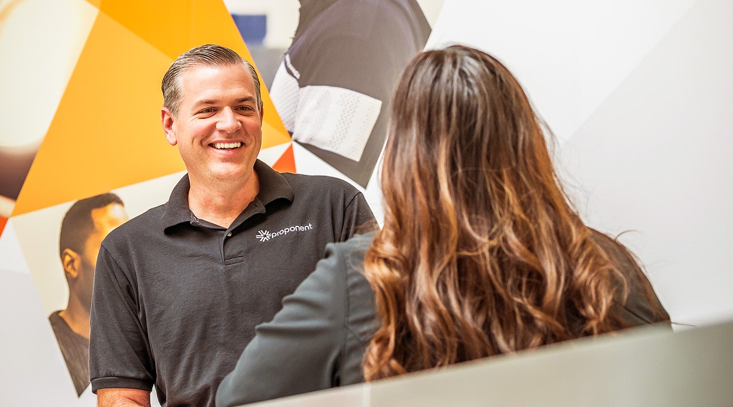 A man and a woman talk, with the man wearing a black, Proponent branded shirt