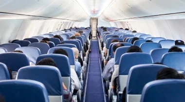 Well lit, interior shot of an aeroplane, showing two banks of seats in rows of three. Blue carpet and seat head rests.