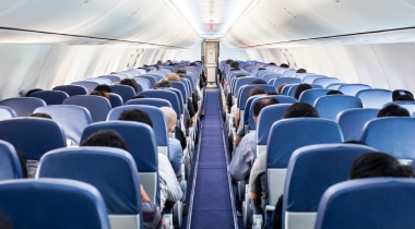 Well lit, interior shot of an aeroplane, showing two banks of seats in rows of three. Blue carpet and seat head rests.