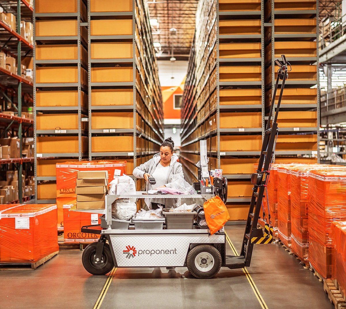 A woman scans an item in a warehouse, on top of a Proponent branded cart.