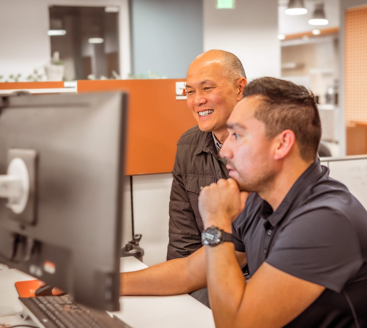 Two men sit at a computer, one is smiling and the other focussing on what's on screen