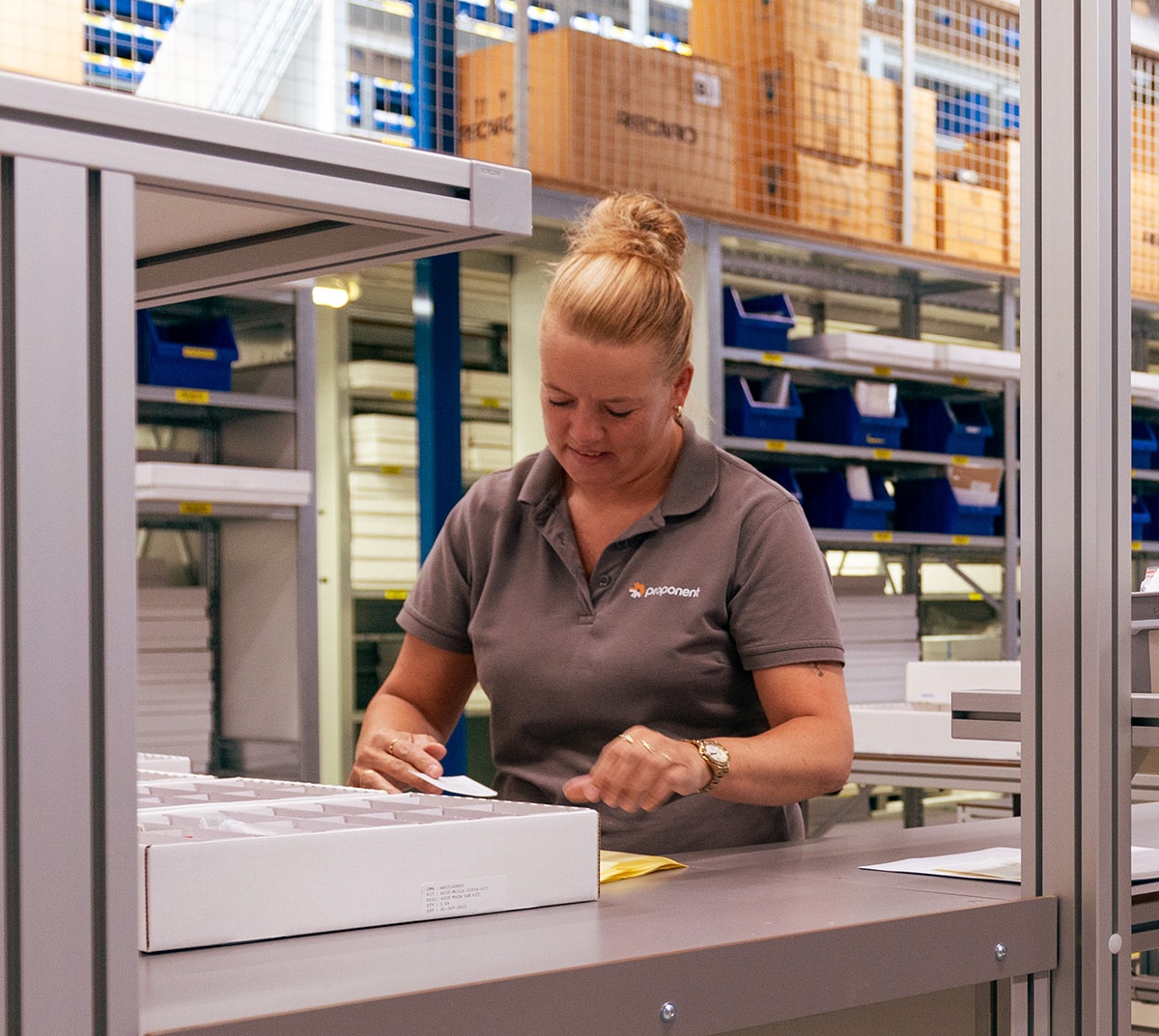 A woman in a warehouse, inspects a label, wearing a grey Proponent branded shirt