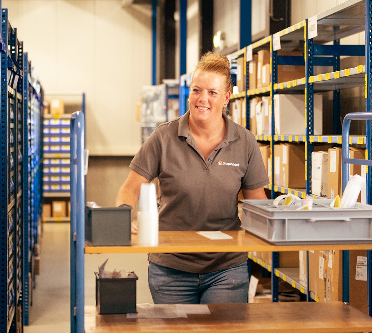 A smiling woman stands behind a set of shelves in a warehouse, wearing a grey Proponent branded shirt