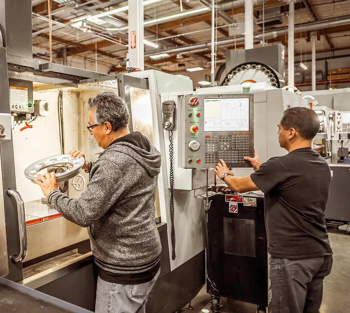 Closeup shot of two men working at a machine. The man on the left is inspecting a part and the man on the right is using the control panel